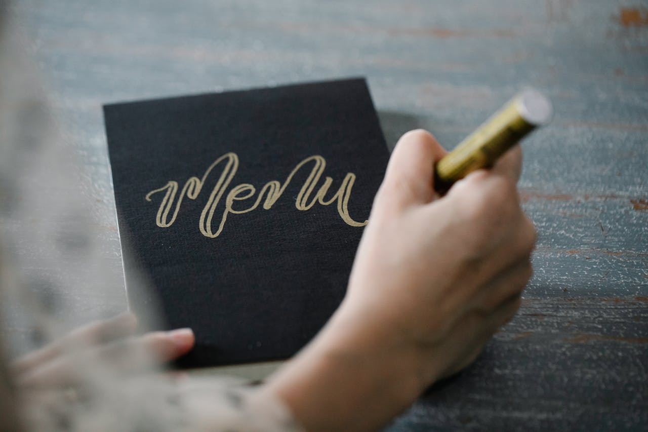 Close-up of a person writing 'Menu' in gold calligraphy on black cardstock with a gold marker.